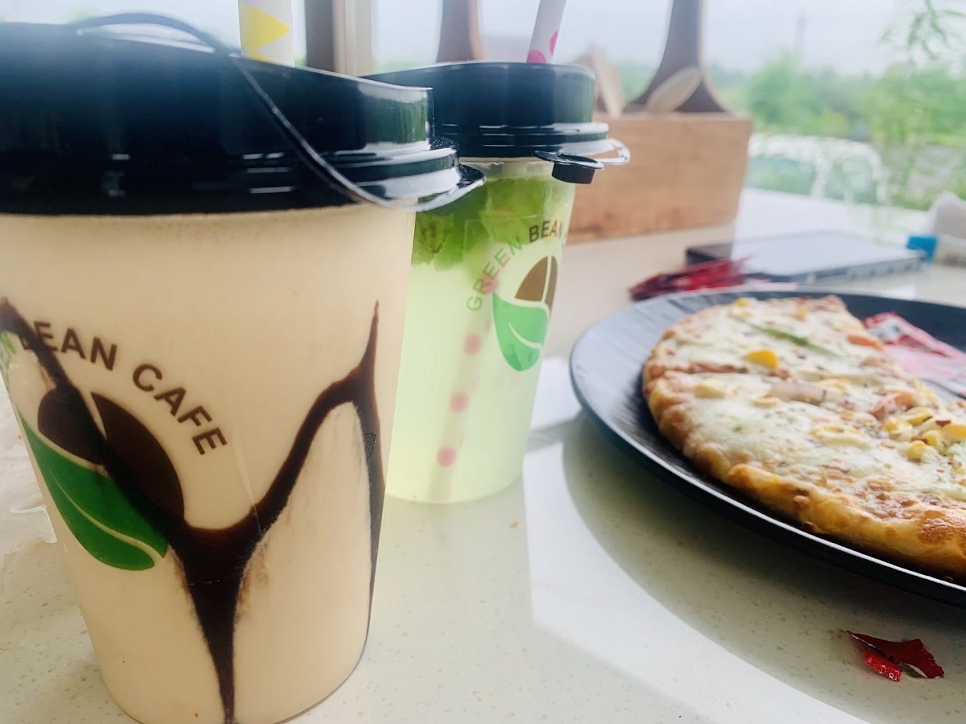 A frappé and a cooler in Green Bean Cafe cups, with a pizza, the garden behind